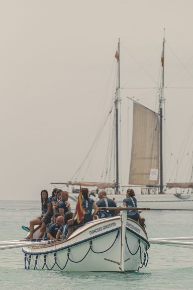 Grupo de personas en un barco, con un velero en el fondo y aguas tranquilas.