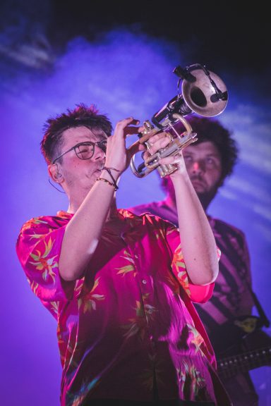 Músico tocando la trompeta, vestido con una camisa colorida, con fondo iluminado.